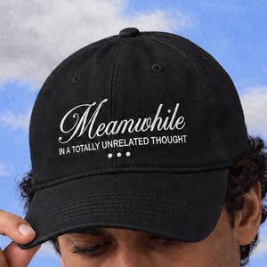May include: A black baseball cap featuring white embroidered text that reads "Meanwhile" in a cursive script, followed by "IN A TOTALLY UNRELATED THOUGHT" in a clean sans-serif font, with three small dots centered below the text.