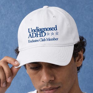 Könnte beinhalten: Eine weiße Baseballkappe mit dem blauen Schriftzug Undiagnosed ADHD Exclusive Club Member und drei blauen Sternumrissen. Die Kappe hat eine strukturierte Krone, einen gebogenen Schirm und einen Knopf an der Oberseite für ein schlichtes Design.