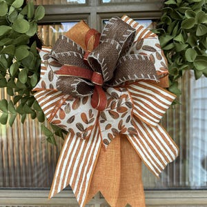 May include: A decorative bow featuring a mix of ribbons in brown, orange, and white. The design includes solid orange fabric, white ribbons with brown leaf patterns, white ribbons with orange stripes, and dark brown textured ribbons with white stitching.