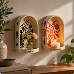 May include: Two arched cream-colored ceramic wall niches. The left niche features black botanical illustrations and the word Faith, holding a small glass vase with flowers. The right niche has red pomegranate designs and the word Blessed, holding a lit tea light candle.