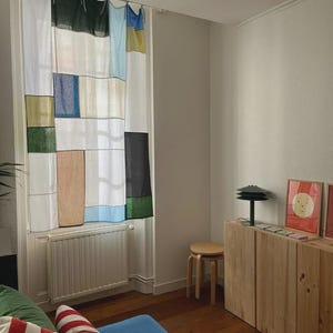 Könnte beinhalten: Ein Fenstervorhang mit einem Patchwork-Design aus rechteckigen Feldern in Weiß, Schwarz, Dunkelgrün, Hellblau, Beige und Gelb. Der Vorhang ist mit Stoffbändern an einer Stange befestigt und hängt vor einem Fenster über einem weißen Heizkörper.