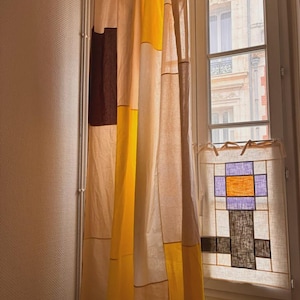 Könnte beinhalten: Ein langer Vorhang mit einem Patchwork-Design aus cremefarbenen, gelben und dunkelbraunen Stoffbahnen. Daneben zeigt ein kleinerer Fensterbehang ein geometrisches Muster mit einem zentralen orangefarbenen Quadrat, violetten Rändern und einem dunkelbraunen Sockel.