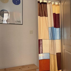 Pode incluir: Uma cortina de porta com um design de patchwork em blocos de cores nos tons creme, bordô, azul claro e amarelo mostarda. A cortina é suspensa por alças de tecido no topo e cobre a porta, exibindo um padrão de grade retangular.