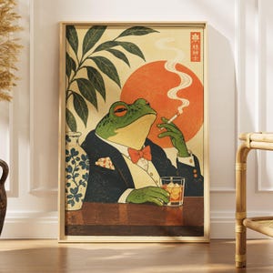 May include: A rectangular wall art print featuring a green frog dressed in a black tuxedo with an orange bowtie, holding a lit cigarette and a glass of whiskey. A large orange sun, a blue and white patterned vase, and green leaves are visible.