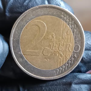 Peut inclure: Une pièce de deux euros au design bimétallique avec un centre doré et un anneau extérieur argenté. La pièce affiche le chiffre 2, le mot EURO et une carte de l'Europe avec des étoiles sur le bord, présentant des signes d'usure et des rayures.