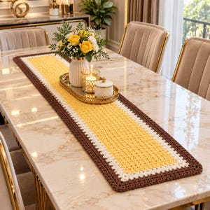 Puede incluir: Un camino de mesa rectangular de ganchillo con un panel central amarillo, un borde blanco y un contorno marrón oscuro. El camino de mesa está sobre una mesa de mármol y sostiene un jarrón blanco con flores amarillas y una bandeja dorada.