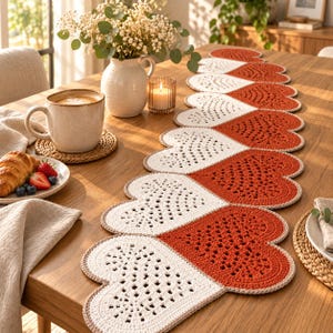Pode incluir: Um longo caminho de mesa decorativo feito de motivos de crochê em forma de coração em cores alternadas branco e laranja queimado. Cada coração apresenta um intrincado padrão de renda aberta com uma borda sólida, disposto em corrente.