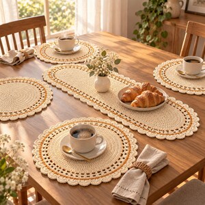 Puede incluir: Un juego de manteles individuales de ganchillo color crema y un camino de mesa ovalado a juego con bordes festoneados y finos detalles dorados. La mesa está puesta con tazas de café blancas, cucharas doradas y croissants.