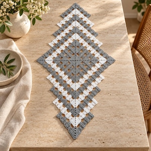 Puede incluir: Un camino de mesa de ganchillo en forma de diamante con un patrón geométrico alterno de hilo blanco y gris. El intrincado diseño calado consta de cuadrados concéntricos que se expanden desde el centro, creando un borde de encaje simétrico.