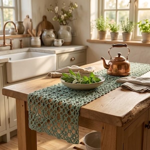 Puede incluir: Un camino de mesa de ganchillo en color verde salvia con un patrón calado sobre una isla de cocina de madera. Una tetera de cobre se sitúa sobre el camino junto a un cuenco de cerámica poco profundo con hierbas frescas y dos servilletas de lino beige.