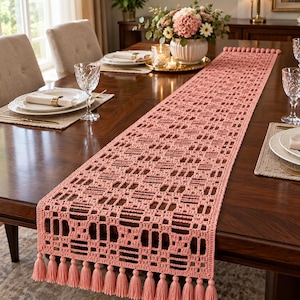 Puede incluir: Un camino de mesa largo y rectangular de ganchillo en color rosa empolvado con un intrincado patrón geométrico calado. El camino está rematado con una hilera de borlas a juego en ambos extremos cortos, creando un acento decorativo texturizado.