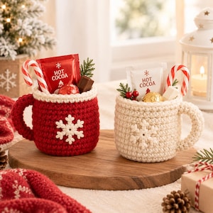 May include: Two crocheted mugs, one red and one cream, each featuring a white snowflake appliqué. The mugs are filled with hot cocoa packets labeled "HOT COCOA," candy canes, and small chocolates, creating a festive holiday display.
