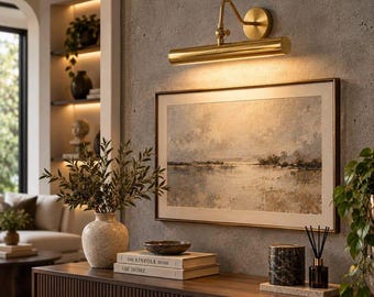 May include: A gold-toned metal picture light fixture mounted on a textured grey wall above a framed landscape painting. The light features a cylindrical shade and an adjustable arm, casting a warm glow downwards onto the artwork below.