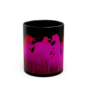 Pode incluir: Uma caneca de cerâmica preta com uma estampa gráfica vibrante em rosa néon de uma banda musical a atuar num palco. O design exibe silhuetas de músicos a tocar instrumentos, criando um efeito visual de alto contraste sobre o fundo escuro.