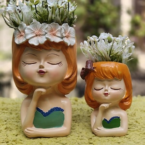May include: Two resin head-shaped planters featuring orange-haired figures with closed eyes and hands resting under their chins. The larger figure wears a white floral crown, while the smaller has a brown hair accessory. Both wear green tops.