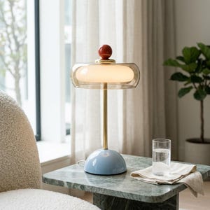 Peut inclure: Une lampe de table avec une base en dôme bleu clair, une tige métallique dorée fine et un large abat-jour en verre teinté ambre transparent entourant un globe intérieur blanc. Un petit embout rouge brillant surmonte l'abat-jour.