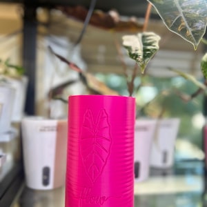 May include: A cylindrical, bright pink plant pot featuring a textured, horizontal ribbed surface. The front of the pot displays an embossed leaf design positioned above the word flow written in a cursive, script-style font.