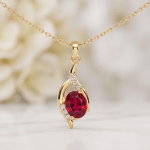 May include: A gold pendant necklace featuring a central oval-cut deep red gemstone surrounded by a swirling gold frame accented with six small round clear stones. The pendant hangs from a delicate gold chain with a polished metallic finish.
