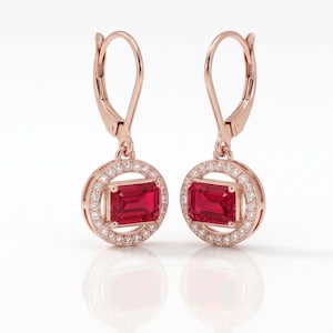 May include: A pair of rose gold drop earrings featuring a central emerald-cut red gemstone surrounded by a halo of small white stones. The earrings are suspended from a lever-back mechanism, showcasing a circular frame design with a vibrant red center.