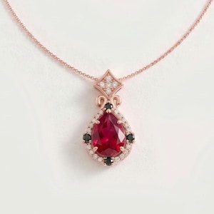 May include: A rose gold necklace featuring a pear-shaped red gemstone pendant surrounded by a halo of small white diamonds and four black stones. The pendant is topped with a diamond-encrusted geometric bail and hangs from a thin chain.
