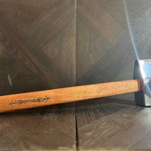 Könnte beinhalten: Ein Hammer mit einem polierten, reflektierenden Metallkopf und einem glatten, hellbraunen Holzgriff. Der Griff weist eine schwarze, lineare, pfeilartige dekorative Gravur auf, die entlang der Seite in der Nähe des Werkzeugendes verläuft.