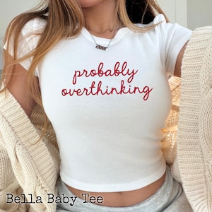 May include: A white short-sleeved baby tee featuring the red cursive text probably overthinking printed on the centre of the chest. The shirt has a crew neckline and a fitted silhouette, shown layered under a cream-coloured cable knit cardigan.