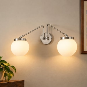 May include: A double-arm wall sconce featuring a polished chrome finish and two spherical, frosted white glass globes. The fixture is mounted to a circular chrome base, with adjustable metal arms extending outward to support the glowing light bulbs.