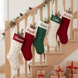 May include: Five cable-knit Christmas stockings hanging in a row, featuring alternating red, green, and cream colours. Each stocking has a contrasting cuff embroidered with the names Sloane, Wyatt, Nina, Cole, and Marley, filled with pine greenery.