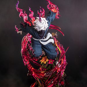 Puede incluir: Una figura detallada de estilo anime de un personaje con cabello blanco y máscara negra, vistiendo un traje azul oscuro con una faja blanca. La figura está rodeada de efectos de energía translúcidos rojos y morados y se apoya sobre una base con flores.