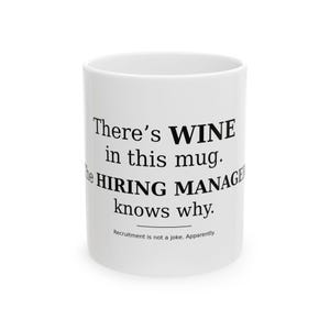 May include: A white ceramic mug featuring black text that reads: There's WINE in this mug. The HIRING MANAGER knows why. Recruitment is not a joke. Apparently. The mug has a cylindrical shape with a smooth finish and a standard handle on the side.