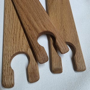 Può includere: Tre lunghe strisce di legno piatte di colore marrone chiaro con una venatura del legno distinta. Ogni striscia presenta un ritaglio circolare a un'estremità, creando un'apertura a forma di U progettata per sostenere un oggetto.