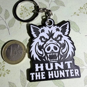 Può includere: Un portachiavi bianco e nero raffigurante una testa di cinghiale stilizzata con la scritta "HUNT THE HUNTER" posizionata sotto. Il portachiavi è mostrato accanto a una moneta da 1 euro per scala, dal diametro di 23,25 millimetri.