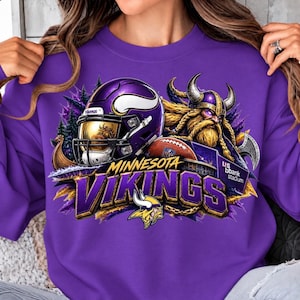 Può includere: Una felpa girocollo viola con una grafica raffigurante un casco da football dei Minnesota Vikings, un guerriero vichingo, un pallone da football e il testo Minnesota Vikings e U.S. Bank Stadium. Il design include accenti viola e oro e immagini di boschi.