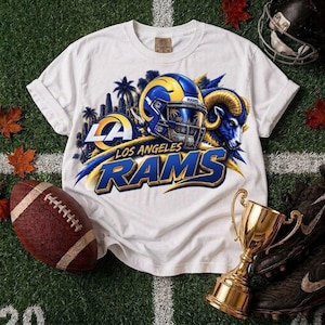 Puede incluir: Una camiseta blanca de manga corta con un gráfico de un casco de fútbol americano azul y amarillo, una cabeza de carnero y el texto Los Angeles Rams. El diseño incluye palmeras y el logotipo LA, impreso en la parte frontal de la prenda.