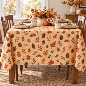 May include: A rectangular tablecloth with a light beige background featuring a repeating autumn-themed pattern of orange and brown maple leaves, pinecones, acorns, red mushrooms, and small animal paw prints, draped over a wooden dining table.