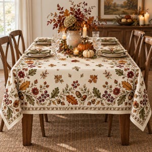 May include: A rectangular tablecloth featuring a cream-colored base with a printed border of autumn leaves, acorns, and red berries. The design includes oak leaves in shades of orange, brown, and green, arranged in a repeating botanical pattern.