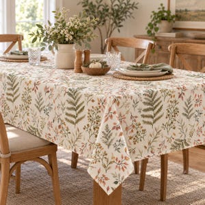 May include: A rectangular tablecloth featuring a repeating pattern of botanical illustrations including ferns and small leaves in shades of green, brown, and orange on a cream background, draped over a wooden dining table with place settings.