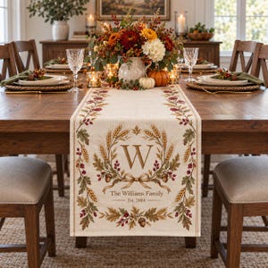 May include: A beige fabric table runner featuring a central wreath design of wheat, oak leaves, and acorns. The letter W is centered within the wreath, with the text The Williams Family and Est. 2004 printed below. The border has a repeating botanical pattern.