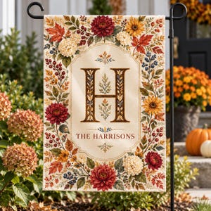 May include: A rectangular garden flag featuring a cream-colored center with a large brown letter H and the text THE HARRISONS. The border is decorated with a vibrant autumn-themed floral wreath of red dahlias, yellow sunflowers, and berries.