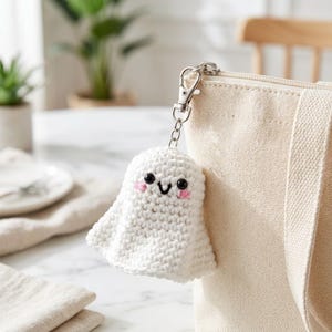 May include: A white crocheted ghost keychain with black bead eyes, a stitched black mouth, and pink embroidered cheeks. The ghost is attached to a silver-toned metal lobster clasp and chain, which is clipped onto a beige canvas tote bag.