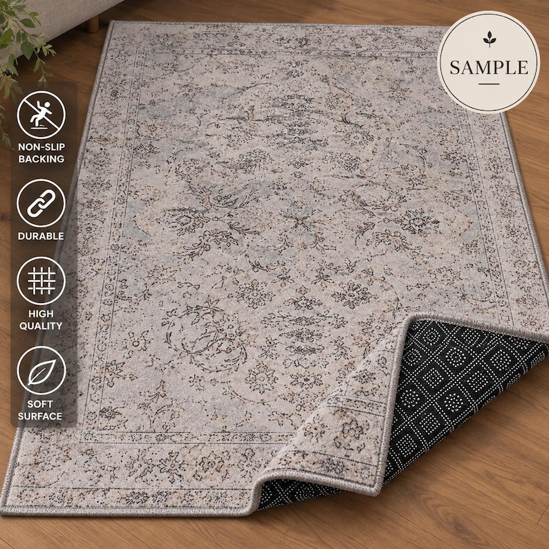 K&ouml;nnte beinhalten: Ein rechteckiger Teppich mit einem hellgrauen und beigen Blumenmuster und einer dekorativen Bord&uuml;re. Der Teppich ist an einer Ecke umgeschlagen und zeigt eine schwarze, rutschfeste Unterseite mit wei&szlig;en Punkten. Der Text SAMPLE ist zu sehen.