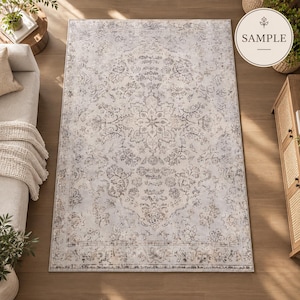 K&ouml;nnte beinhalten: Ein rechteckiger Teppich mit hellgrauer und cremefarbener Basis, verziert mit einem verblassten, komplexen floralen Medaillonmuster in Dunkelgrau und Beige. Das Design umfasst eine dekorative Bord&uuml;re und das Wort SAMPLE in einem runden Abzeichen.