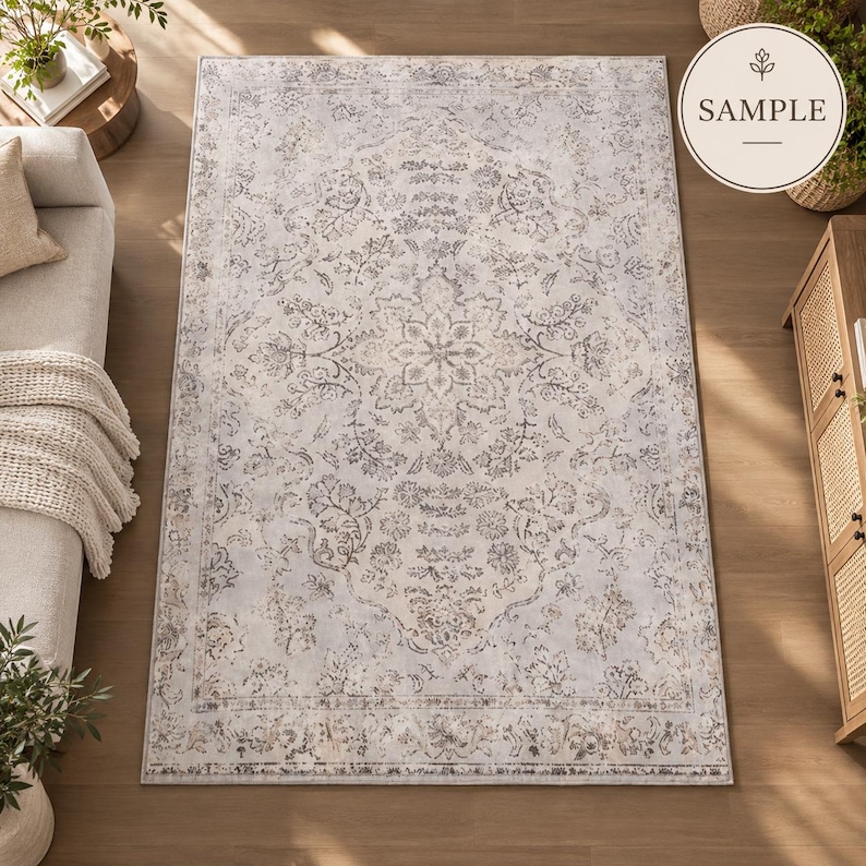 K&ouml;nnte beinhalten: Ein rechteckiger Teppich mit hellgrauer und cremefarbener Basis, verziert mit einem verblassten, komplexen floralen Medaillonmuster in Dunkelgrau und Beige. Das Design umfasst eine dekorative Bord&uuml;re und das Wort SAMPLE in einem runden Abzeichen.