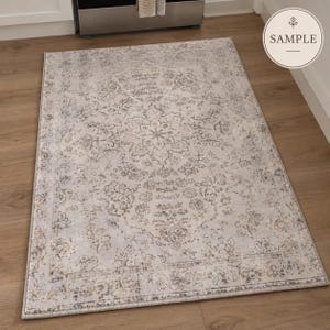 K&ouml;nnte beinhalten: Ein rechteckiger Teppich mit einem verblassten, abgenutzten Muster in den Farben Hellgrau, Beige und Creme. Das Design umfasst ein zentrales Blumenmedaillon, das von komplizierten botanischen Motiven und einem dekorativen Rahmen umgeben ist.