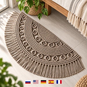 May include: A semi-circular macrame rug in a neutral beige tone, featuring a central woven pattern, concentric rows of knotted cord, and a decorative fringe border along the curved edge. The design consists of repeating circular motifs.