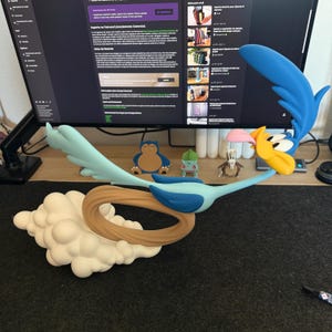 May include: A Road Runner figurine in a dynamic running pose, featuring a bright blue body, yellow beak, and pink tongue. The character is attached to a brown circular motion blur effect, which is mounted on a white, cloud-like base structure.