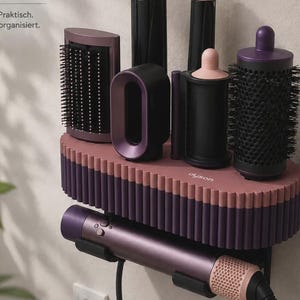 Peut inclure: Un support mural de rangement pour outils de coiffure avec une base cannelée mauve et une bordure violet foncé. L'unité contient un sèche-cheveux violet et divers accessoires de coiffage noirs et violets, avec le texte 'dyson' visible.