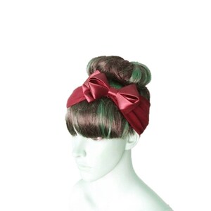 May include: A burgundy satin headband with a large bow. The headband is worn on a mannequin with brown and green hair.