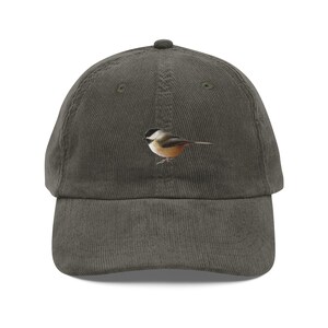 Op de afbeelding: Een donker olijfgroene corduroy baseballpet met een geborduurde afbeelding van een kleine vogel met een zwarte kop, witte wangen en een bruinachtig lichaam, gecentreerd op het voorpaneel van de pet boven de gebogen klep.