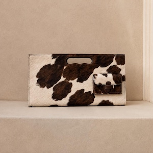 Könnte beinhalten: Eine rechteckige Clutch mit braun-weißem Kuhfellmuster. Die Tasche verfügt über einen ausgeschnittenen Griff in der oberen Mitte und eine kleine, aufgesetzte Außentasche auf der rechten Vorderseite mit Klappverschluss und dunkelbraunem Riemen.